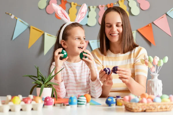 Indoor shot of charming family preparing together for holiday, mother and her child painting Easter eggs, cute girl wearing bunny ears having amazed expression.