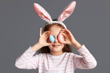 Mutlu Paskalyalar. Evde, tavşan kulağı takan, Paskalya yumurtalarını tutan, gözlerini testislerle kaplayan, eğlenen, gri arka planda soyutlanmış küçük bir kız resmi.