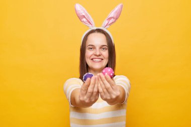 Tavşan kulağı takan, gülümseyen, iki el boyalı Paskalya yumurtası tutan, kameraya bakan ve sarı arka planda izole edilmiş mutluluğu ifade eden bir kadın resmi.