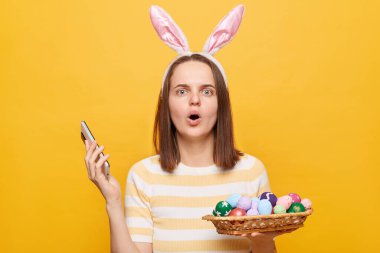 Kapalı alanda, tavşan kulağı takan, cep telefonunda ve sepette Paskalya yumurtası tutan, büyük gözlerle kameraya bakan ve sarı arka planda izole edilmiş ağzı açık bir kadının görüntüsü.