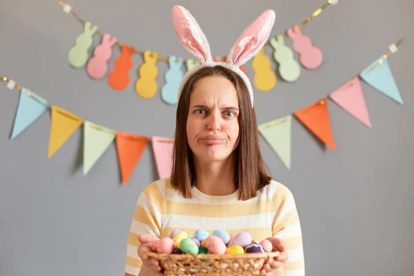 Indoor shot of disappointed sad upset woman wearing rabbit ears holding colorful Easter eggs in basket, looking at camera with sorrow, celebrating holiday alone, isolated on gray decorated background