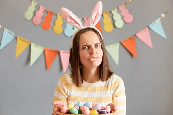 Image of nervous sad depressed woman wearing rabbit ears holding multicolored Easter eggs isolated on gray decorated background, waiting for guests, biting her lips