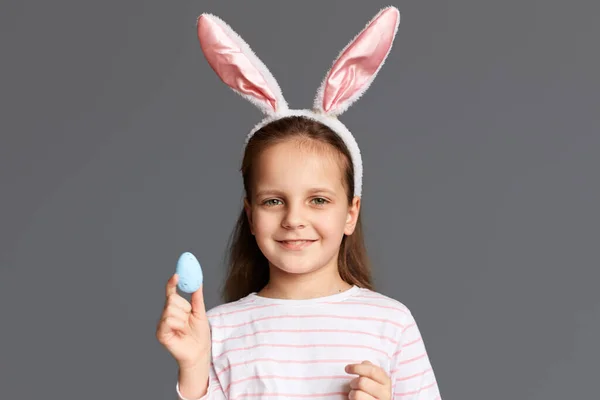 照片上 可爱可爱的 积极的小女孩 戴着小兔子耳朵 手里拿着她自己画的复活节彩蛋 准备度假 面带微笑 在灰色的背景下孤立无援 — 图库照片
