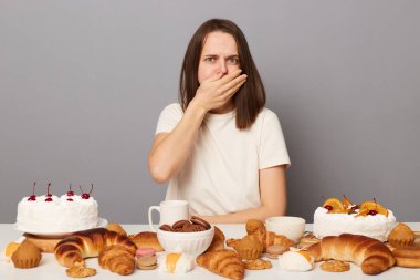 Aşırı yemek yiyen, kahverengi saçlı, gri arka planda tek başına oturan, bol bol tatlı fırın yiyen, kötü hisseden, mide bulantısı çeken, palmiye ile ağzını kapatan genç bir kadının kapalı çekimleri..