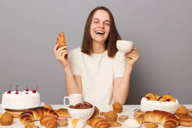 Kapalı alanda gülümseyen, kahverengi saçlı, gri arka planda izole bir şekilde oturan, ayçöreğiyle çay içen, akşam yemeğinin tadını çıkaran, mutlu bir şekilde gülen kadının fotoğrafı..