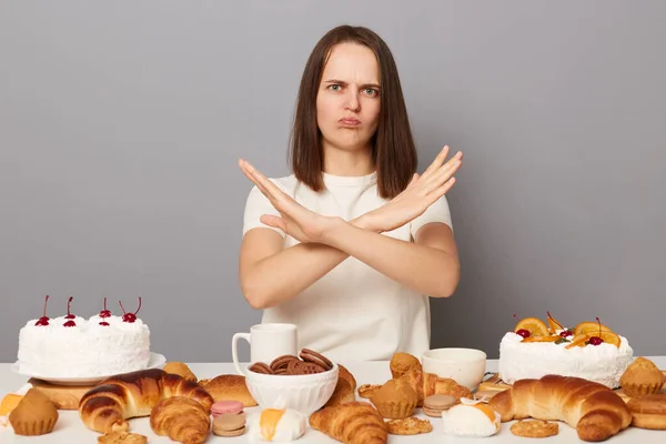 Portrait of sad serious woman with brown hair sitting at table full of pastry, showing no way gesture, crossed arms, saying do not eat unhealthy bakery isolated over gray background.