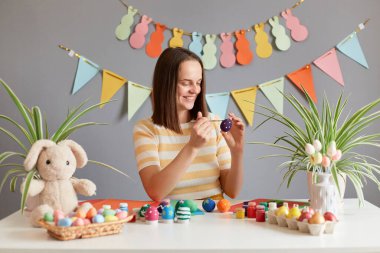 Siyah düz saçlı, gülümseyen bir kadının portresi çizgili gömlek giyiyor. Paskalya yumurtalarını boyuyor. Tatile hazırlanıyor.