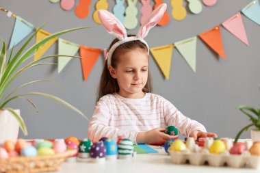 Kapalı alanda, tavşan kulağı takan, paskalya için masa başında oturan, süsleri ve boyalı yumurtaları olan, gri duvara karşı hazırlanan koyu renk saçlı küçük bir kız resmi..