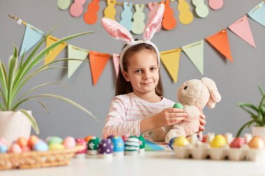 Kapalı alanda, tavşan kulağı takan, paskalya için hazırlanan, süslemeli, boyalı yumurtalı ve tavşan oyuncağıyla oynayan küçük sevimli bir kız resmi..