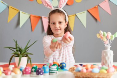 Kapalı alanda, tavşan kulağı takan, renkli yumurtaları gösteren, bahar tatilinin tadını çıkaran, gri duvarlı Paskalya 'ya hazırlanan güzel bir kız resmi..