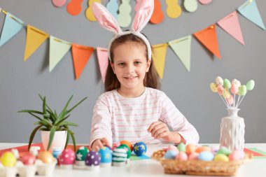 Mutlu Paskalyalar. Pozitif iyimser gülümseyen, tavşan kulağı takan küçük bir kızın fotoğrafı. Paskalya için hazırlanıyor. Gri duvarda süslerle duruyor, kameraya bakıyor..
