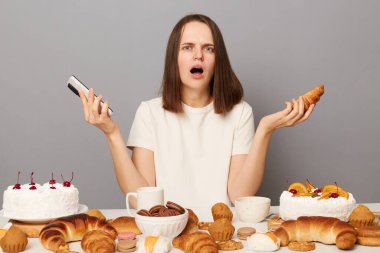 Kahverengi saçlı, beyaz tişörtlü, gri arka planda yalnız başına oturan öfkeli kadın, günde ne kadar kalori yediğine şaşırdı, tatlı ve akıllı telefon tutuyor, diyeti yok..