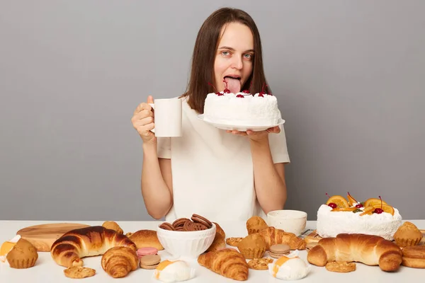 Photo of hungry woman with brown hair wearing white T-shirt sitting at table isolated over gray background, drinking tea and licking cake, looking at camera.
