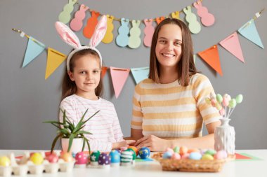 Kapalı alanda anne ve kızının tavşan kulağı taktığı, Paskalya için yumurta boyarken, gri dekore edilmiş duvara bakarken, neşeli bir ifadeyle kameraya bakarken, neşeli bir ruh hali içindeyken..