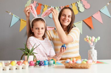 Neşeli insanların bahar tatili boyunca eğlendikleri portre, anne ve kız, çocuğun kulaklarına dokunan kadın yumurta boyuyor gri dekore edilmiş duvarlara karşı Paskalya için hazırlanıyorlar..