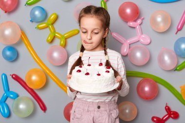 Saçları günlük giysiler giyen, gri duvarda renkli balonlarla dikilen, güzel lezzetli pastaya bakan, dilini gösteren aç küçük bir kızın portresi..