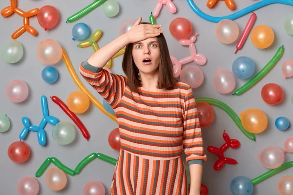 Portrait of shocked sad caucasian woman with brown hair wearing striped dress, showing facepalm gesture, forgot about birthday present, standing around multicolored inflated balloons.