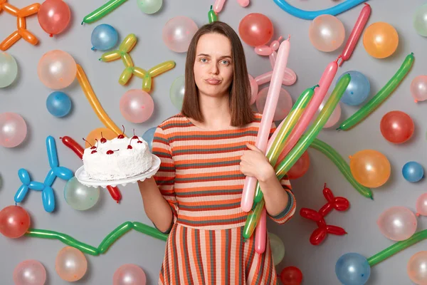 Portrait of sad upset woman with brown hair wearing striped dress standing with cake in hands, waiting her guests, nobody come to party posing around multicolored inflated balloons.