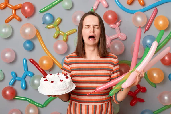 Portrait of despair caucasian woman with brown hair wearing striped dress standing around multicolored inflated balloons, holding cake and crying, feels lonely on her birthday.