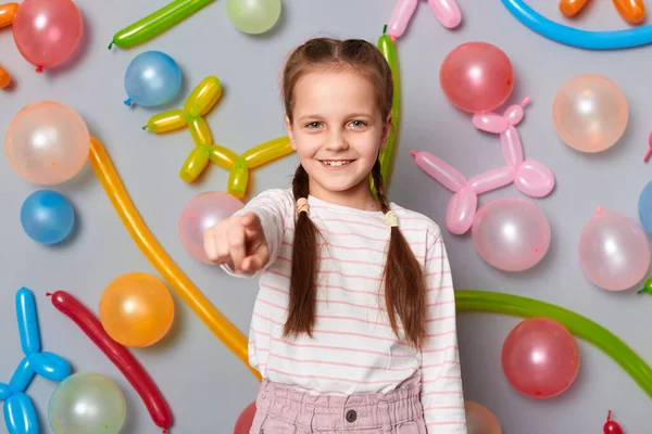 Image of smiling satisfied friendly little girl with braids wearing casual clothing pointing at camera, inviting to her party, posing isolated over gray background with balloons.