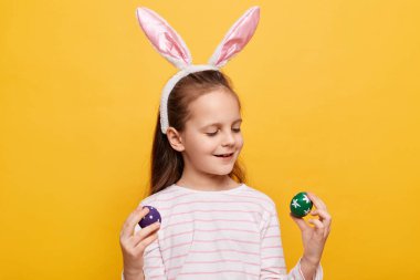 Tavşan kulaklı güzel bir kızın portresi ellerinde renkli yumurtalar, sarı arka planda izole edilmiş gülümsemeli paskalya sembollerine bakıyor..