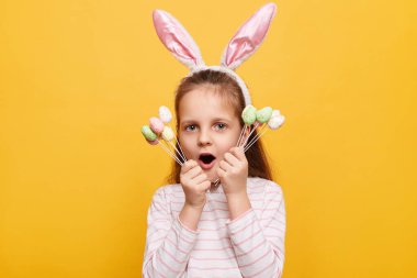 Kafasında tavşan kulakları olan korkmuş küçük bir kızın portresi sarı arka planda izole bir şekilde poz veriyor, elinde pasta çubukları, ağzını açık tutuyor..