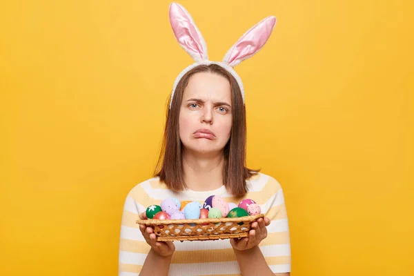 Horizontal shot of sad upset woman wearing bunny ears holding easter eggs isolated over yellow background, looking at camera with pout lips, celebrating holiday alone.