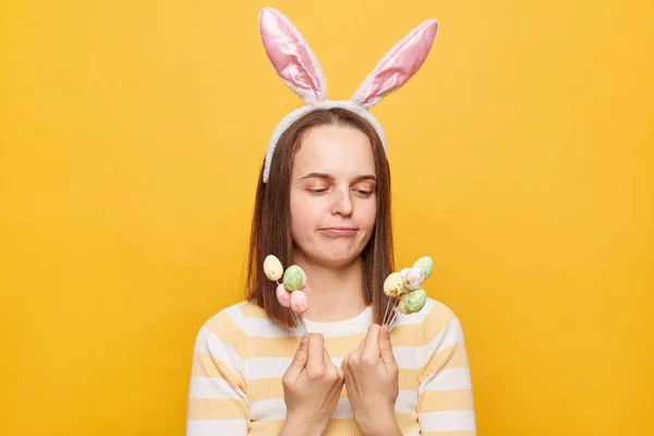 stock image Portrait of confused puzzled woman wearing bunny ears holding easter cake pops isolated over yellow background, deciding eating or not sugary dessert.