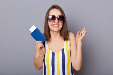 Yüzünde mayo ve güneş gözlüğü olan umut dolu bir kadının portresi parmaklarını çapraz yapıyor, yurtdışına seyahat etmeyi diliyor, elinde pasaport ve biletlerle gri arka planda tek başına duruyor.