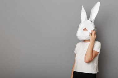 Beyaz t-shirt ve kağıttan tavşan maskesi giymiş, gri arka planda izole bir şekilde duran, akıllı telefondan konuşan, reklam için fotokopi çeken,.