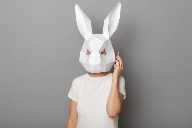 Beyaz t-shirt ve kağıt tavşan maskesi takmış, cep telefonuyla konuşan, kameraya bakan, gri arka planda tek başına duran, tanınmayan bir kadının kapalı çekimleri..