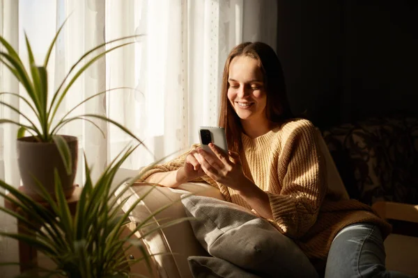 Крытый Снимок Улыбающейся Расслабленной Радостной Женщины Сидящей Диване Используя Мобильный — стоковое фото
