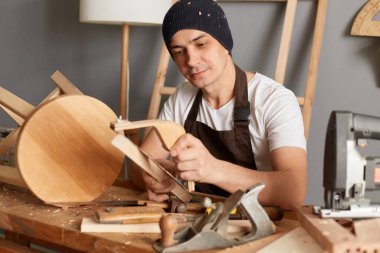 Önlük ve kep takmış, atölyede çalışan, elinde öğütücü tutan, ahşap tahtayı yoğun bakışla öğüten odaklanmış genç marangoz portresi..
