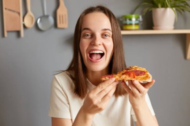 Mutlu, neşeli bir kadının lezzetli bir dilim pizza yerken, fast food atıştırırken, hayret dolu bir suratla kameraya bakarken, mutfakta poz verirken.