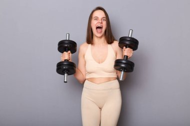 Spor kıyafetleri giyen neşeli, heyecanlı genç bir kadının gri arka planda izole haldeki dambılları tutuşu, ilham verici fiziksel eğitim, çığlıklar..