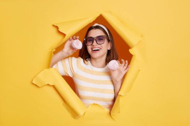 Kapalı alanda, saç bandı ve güneş gözlüğü takan, sarı kağıt delikte duran, lokum tutan, mutlu olan, lezzetli şekerlerden yiyen genç bir kadının fotoğrafı.
