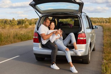 Sevgi dolu bir çiftin arabalarında otururken görüntüsü yolda, erkek ve kadın cep telefonu kullanıyor, selfi çekiyor, birlikte seyahat eden beyaz gömlekler giyiyor..