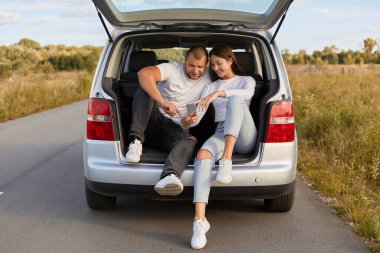 Gülümseyen neşeli bir çiftin görüntüsü yolda arabalarında oturuyor cep telefonu kullanıyor, beyaz tişörtlü bir adam ve kadın otomobille seyahat ediyorlar..