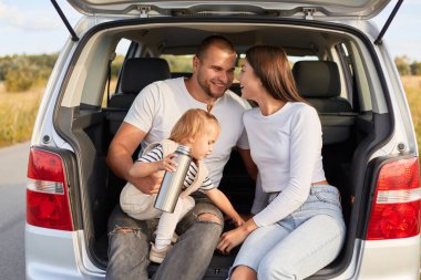 Arabayla gezen, termostan çay ya da kahve içen neşeli aile fotoğrafı. Bir yandan da arabanın bagajında aşk ve mutluluğu ifade ediyor..