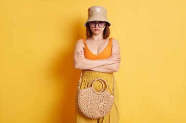 Saman çantasıyla yaz kıyafetleri giyen, sarı arka planda kollarını kavuşturmuş ve dudaklarını büzmüş üzgün genç bir kadının kapalı çekimleri.