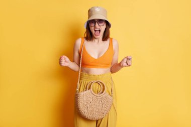 Saldırgan, kahverengi saçlı, yaz kıyafetleri giyen, sarı arka planda izole edilmiş, öfke ve nefret çığlıkları atan, tartışan, saman çantalı genç bir kadının fotoğrafı.