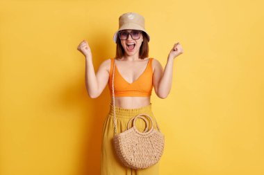 Son derece mutlu genç bir kadının portresi. Yazlık giysiler ve saman çantasıyla sarı arka planda izole bir şekilde duran, yumruklarını sıkan, kazanan, mutluluktan bağıran..