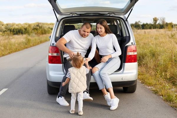 stock image Portrait of happy family having trip around the word, stop to have break and drink tea, sitting in automobile trunk, expressing positive emotions, wearing casual clothing.