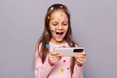 Heyecanlı, kahverengi saçlı, çıkartmalarla kaplı, gri arka planda izole edilmiş, elinde akıllı bir telefonla video oyunu oynayan küçük bir kızın görüntüsü..