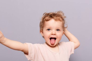 Neşeli gülümseyen çocuk. Kapalı alanda, dilini gösteren küçük bir kız çocuğu, ağzı açık duruyor ve gri arka planda izole bir şekilde dilini dışarı çıkarıyor..