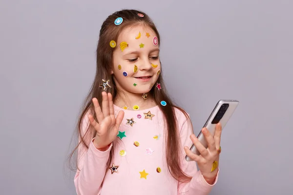 Photo of cute confident little brown haired little girl covered with stickers posing isolated over gray background, having video call, saying hello or bye, waving hand.