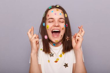 Gri arka planda izole edilmiş komik çıkartmalarla kaplı son derece mutlu bir kadının yatay görüntüsü. Parmaklarını çapraz yapıp, iyi şans ve başarı için dua ediyor..