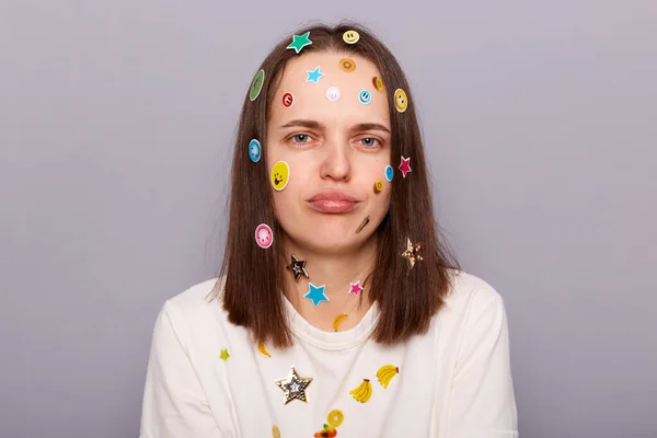 Indoor shot of sad stressed offended disappointed woman covered with funny stickers looking at camera with pout lips, being upset, posing isolated over gray background