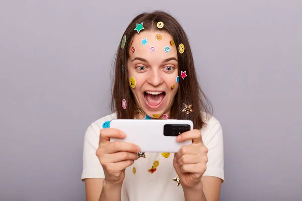 Image of excited woman gamer covered with funny stickers posing isolated over gray background, playing video games, winning level, looking at mobile phone screen.