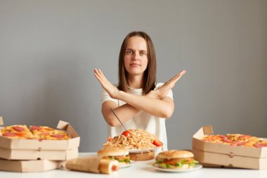 Kahverengi saçlı, beyaz tişörtlü çekici bir kadının portresi gri arka planda yalnız bir masada oturuyor, jest yapmıyor, diyet yapıyor, fast food yapmayı bırakıyor..